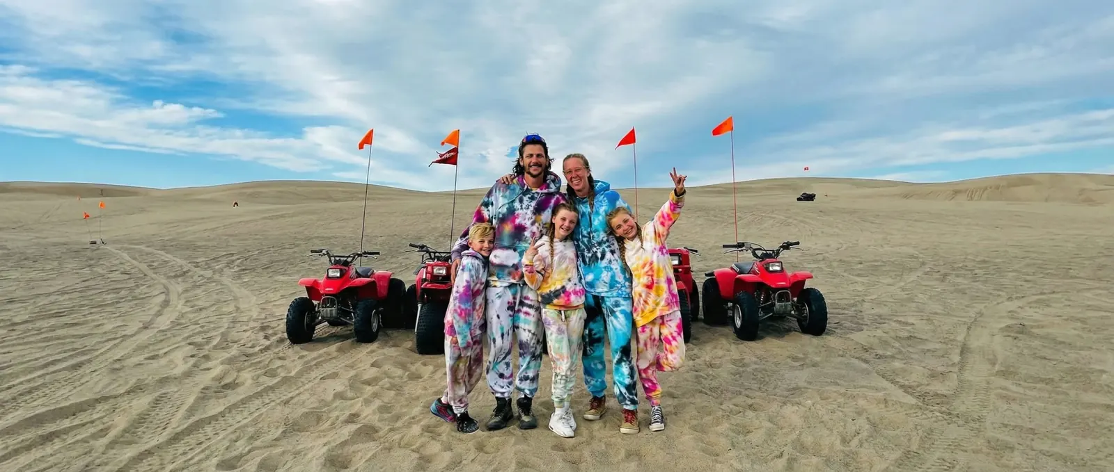 Jake, his wife and their three kids in matching onesies on the sand dunes, flanked by two red ATVs
