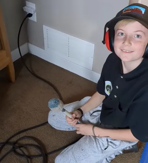 Jake's son in ear protection cleaning a floor vent cover by hand