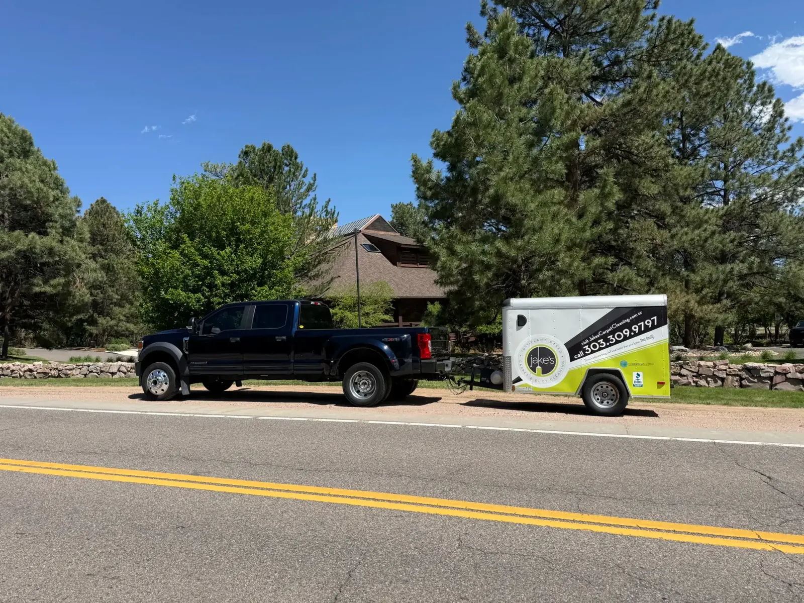 Jake's pickup and branded trailer parked on a pine-lined road outside a mountain home